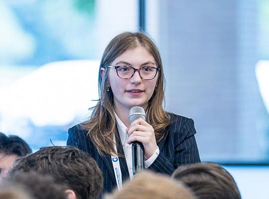 A young woman holds a microphone while speaking in a group.