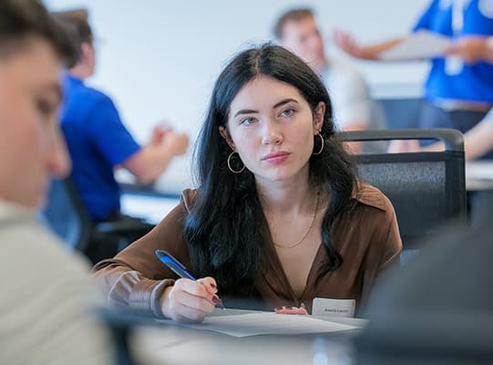 A young woman listens during class while taking notes.